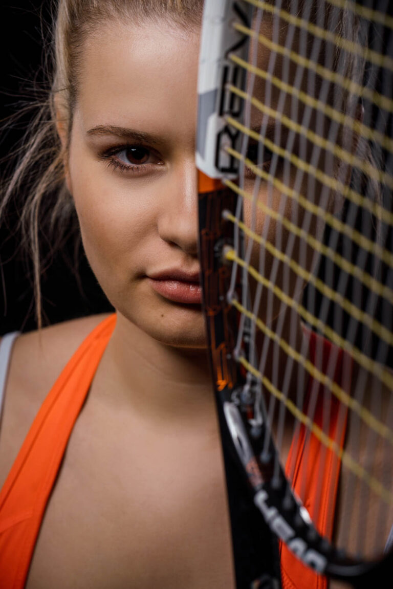 junge Tennisspielerin schaut selbstbewusst in die Kamera mit Schläger vor halbem Gesicht